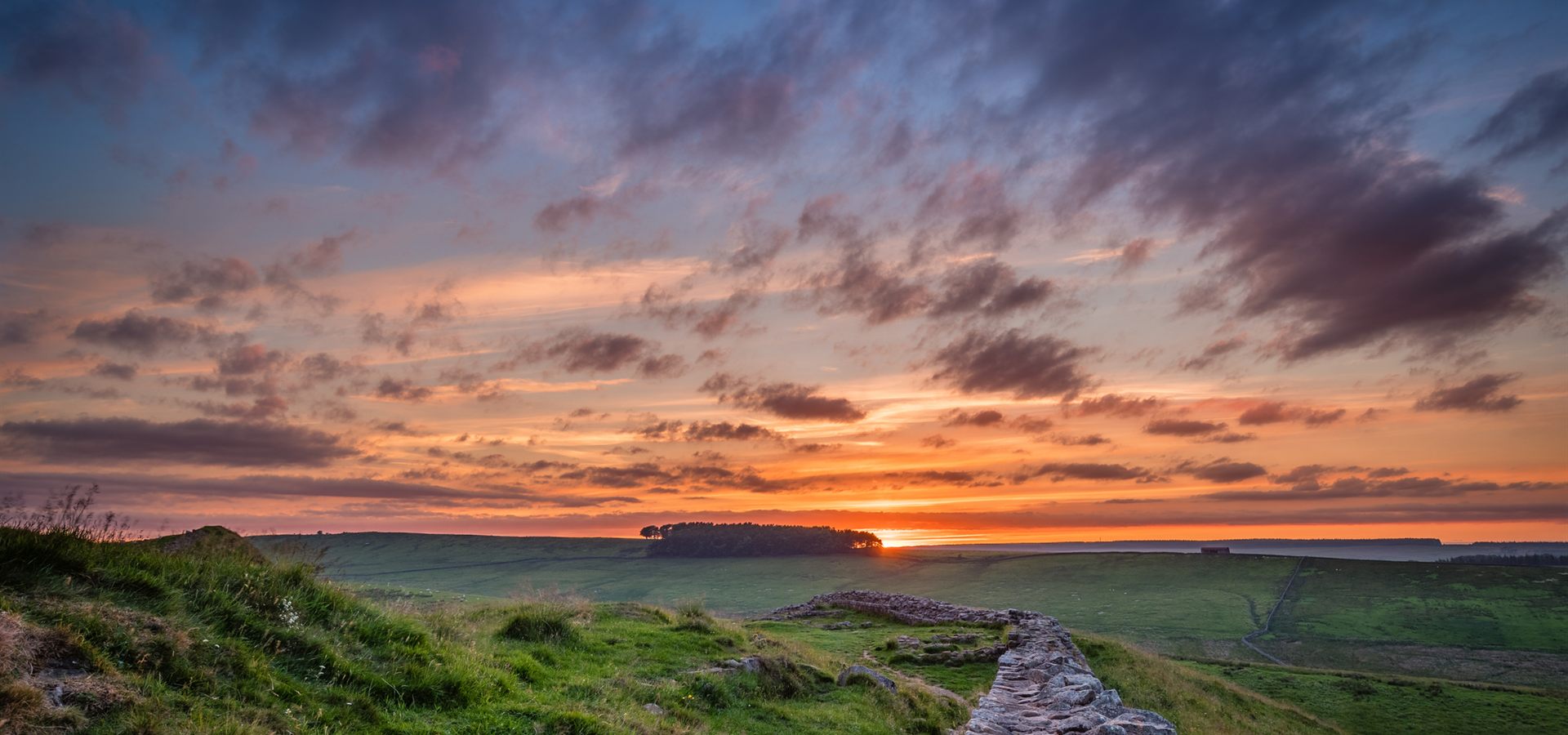 The Hadrian's Wall Path at sunset, the wall running along a grassy ridge above the Northumberland fells.